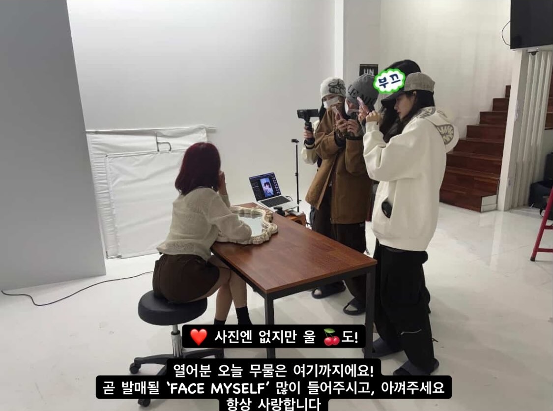 a behind-the-scene photo of face myself photoshoot. jimin is sitting on a chair while the rest of weeekly members (excluding jihan) are standing around her clicking images. there is the text "부끄" on the face of one member.