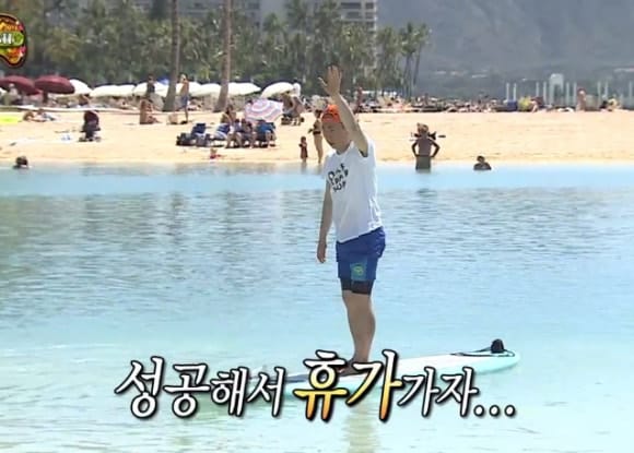 a korean variety show host standing on a surf board in the water next to a beach.